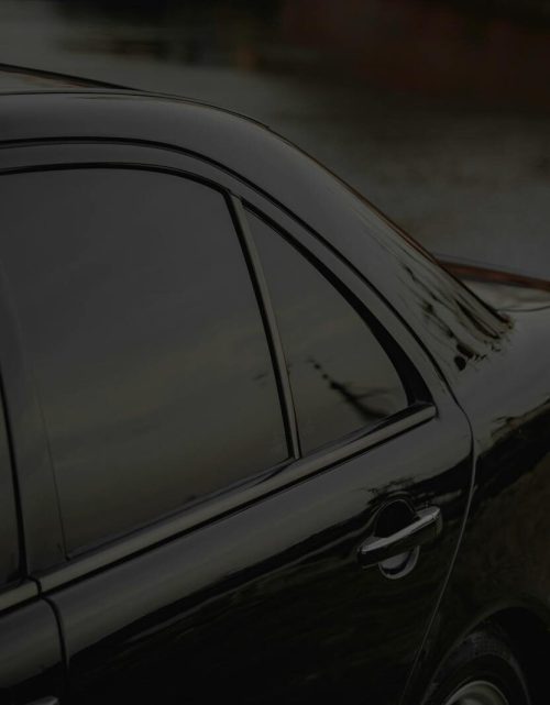 Close-up of a black car's side view with tinted windows by a calm riverbank.